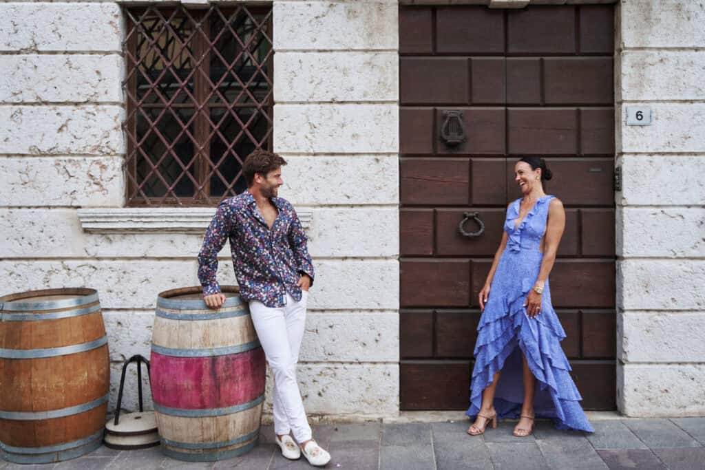 couple smiling and sharing a candid moment in front of a rustic Italian building with wine barrels and a wooden door