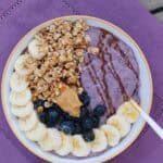 Blueberry chia pudding breakfast bowl topped with banana slices, granola, blueberries, peanut butter, and a drizzle of honey on a purple linen placemat.