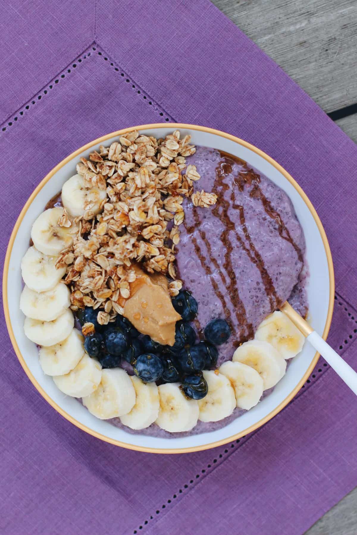 Blueberry chia pudding breakfast bowl topped with banana slices, granola, blueberries, peanut butter, and a drizzle of honey on a purple linen placemat.