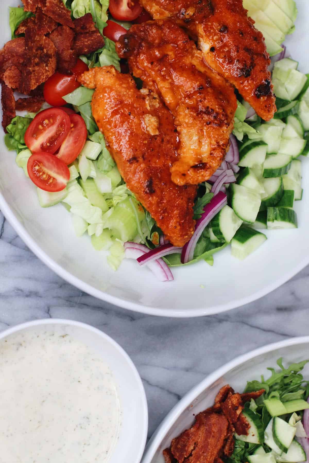 Buffalo chicken salad with homemade ranch dressing, crisp lettuce, cucumbers, cherry tomatoes, red onion, and bacon in a white bowl on a marble surface.