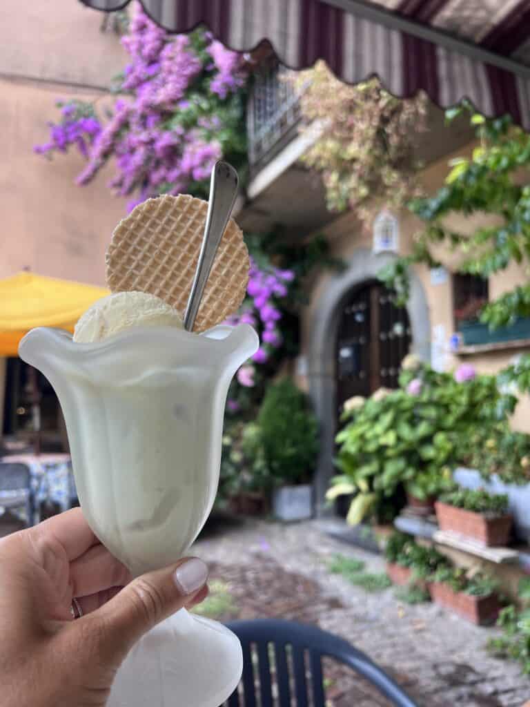 Hand holding a glass of Italian gelato with a wafer and spoon, set against a charming courtyard with blooming flowers and rustic architecture.