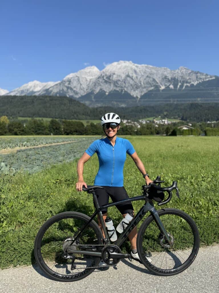 Female cyclist posing with road bike in Innsbruck, Austria, with snow-capped mountains and green farmland in the background.