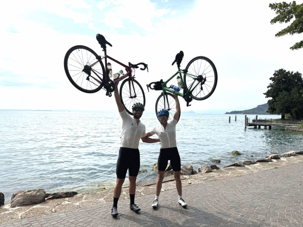 Two cyclists holding their bikes overhead in front of Lake Garda, celebrating at the edge of a cobblestone path by the water.