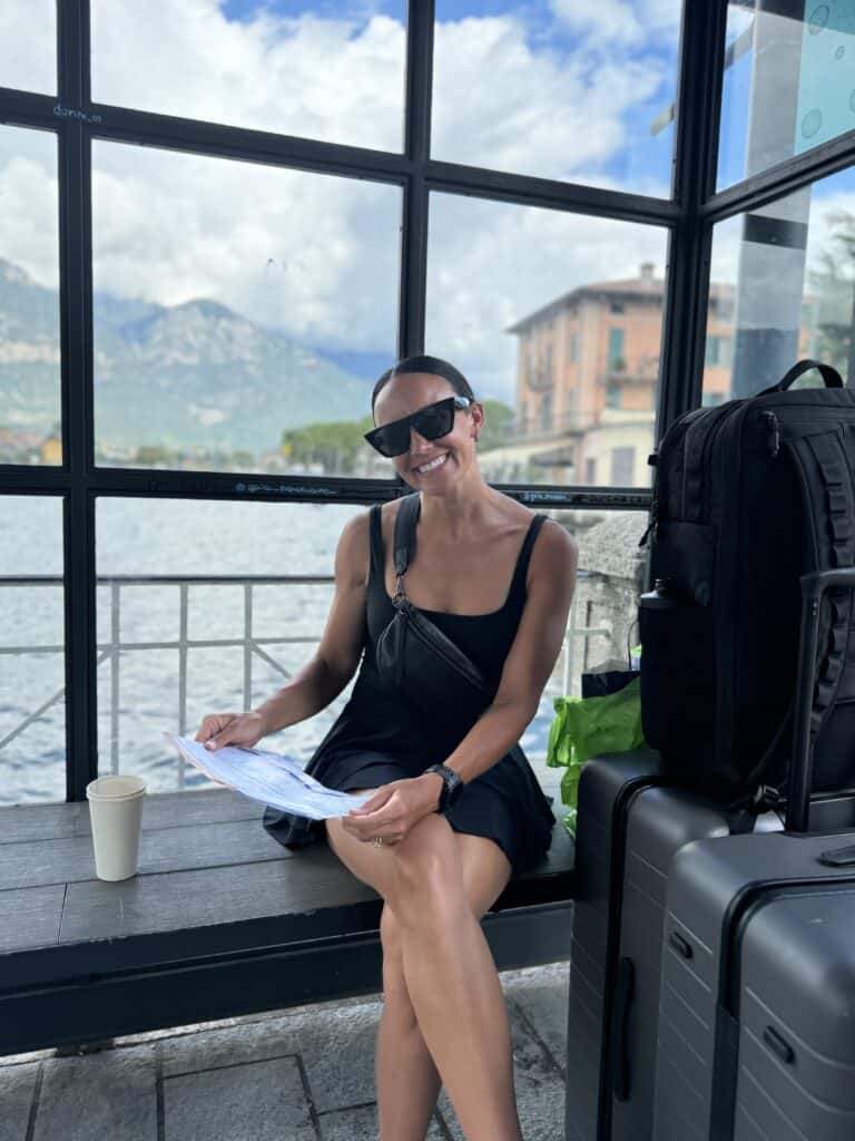 Woman smiling while holding a map at a ferry stop on Lago d'Iseo, with travel bags beside her and mountains in the background.