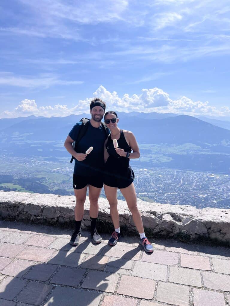 Couple smiling with popsicles at the summit of a hike in Innsbruck, Austria, overlooking the valley and mountains in the background.