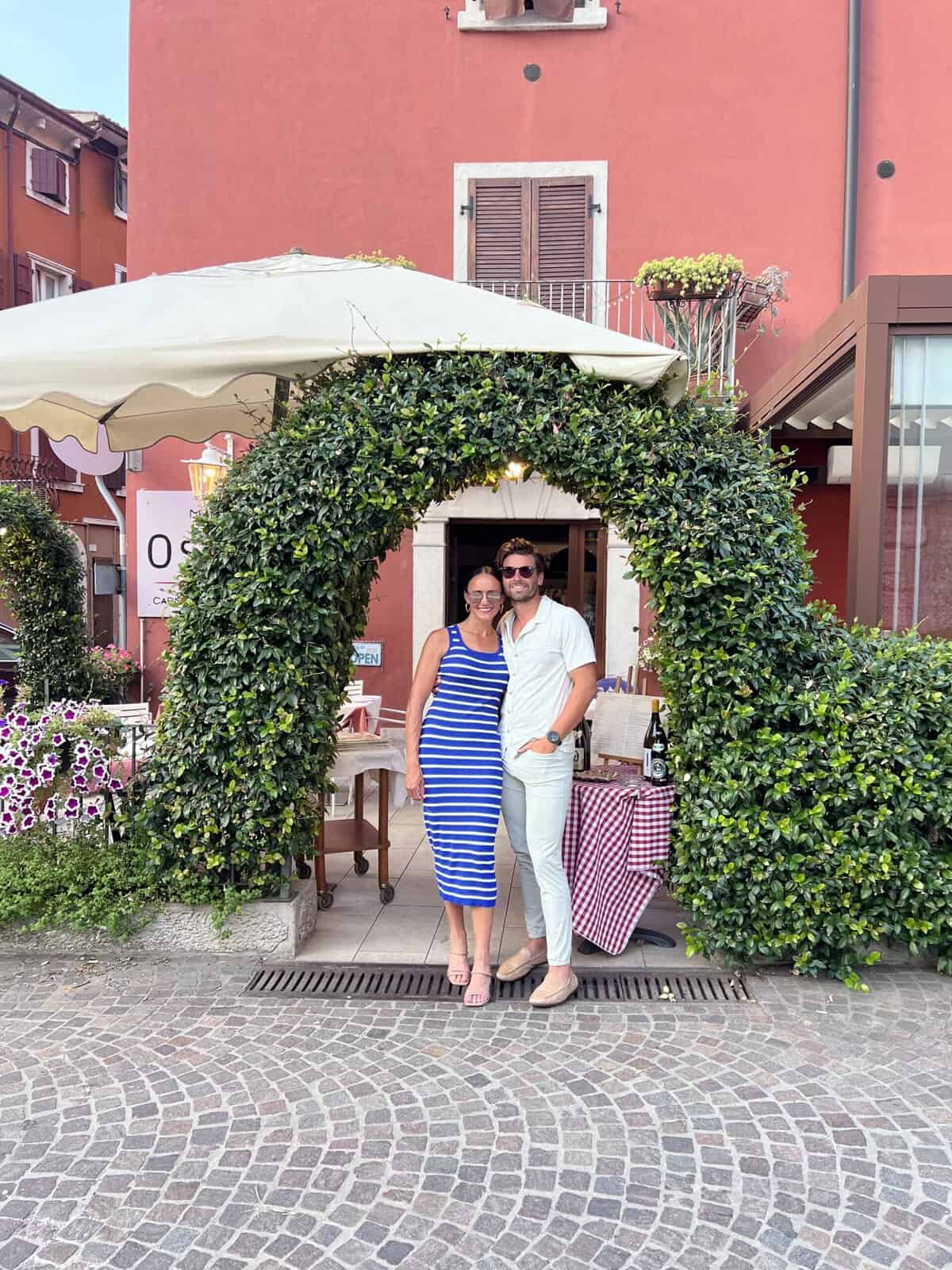 Couple standing under a green ivy archway in front of a charming Italian restaurant with red walls and cobblestone streets.