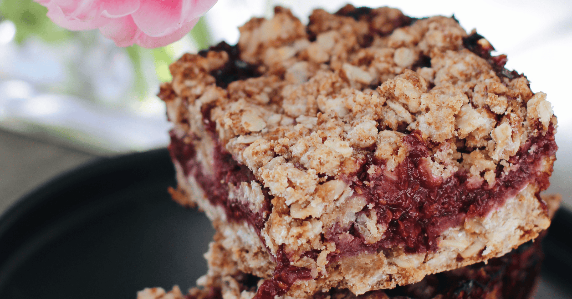 close-up of a gluten-free berry crumble bar stacked on a black plate, with a pink flower in the background