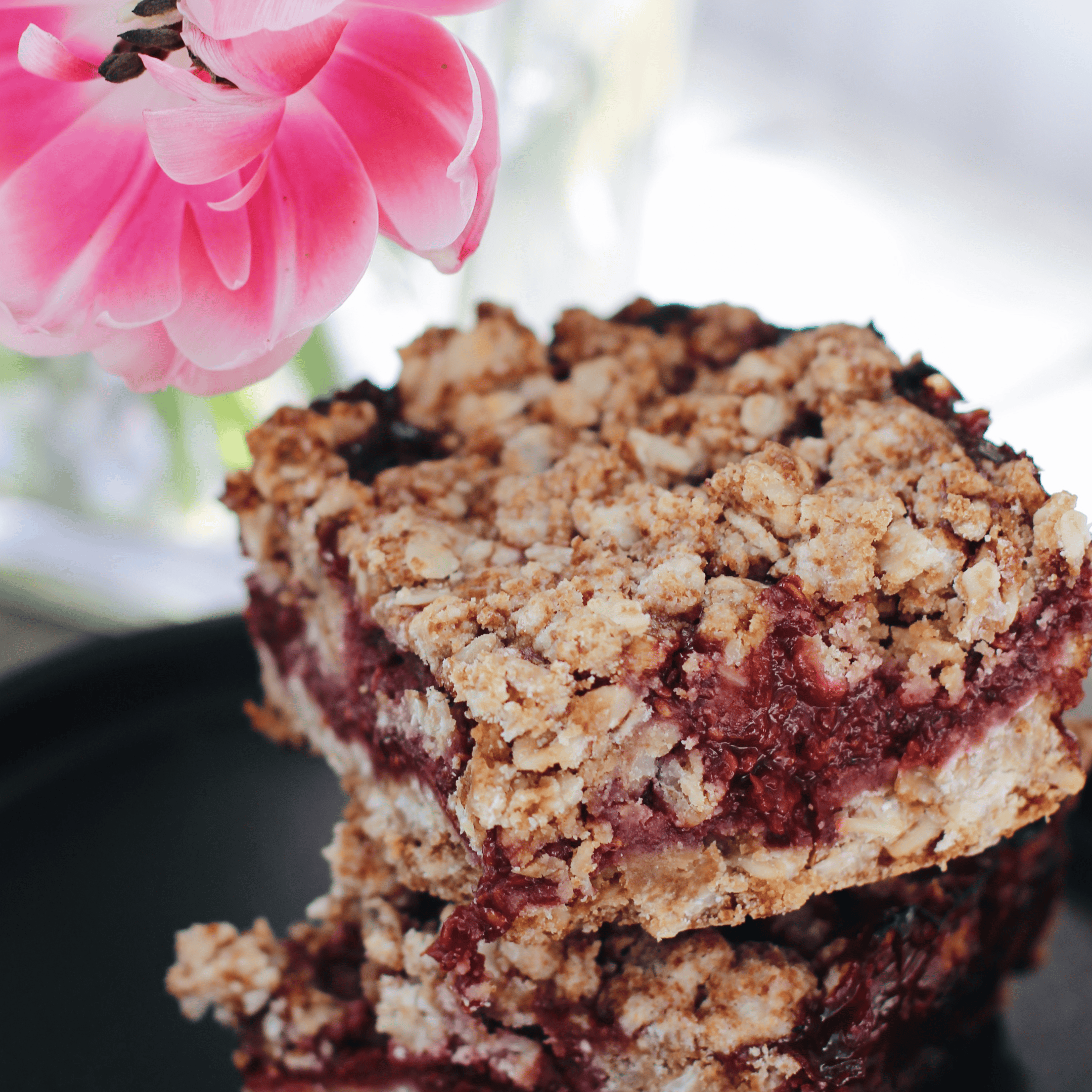 close-up of a gluten-free berry crumble bar stacked on a black plate, with a pink flower in the background