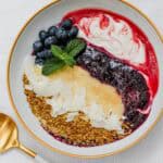 Overhead image of a smoothie yogurt bowl with layered strawberry, blueberry, and pineapple coconut blends, swirled with yogurt and topped with blueberries, bee pollen, coconut, and mint.