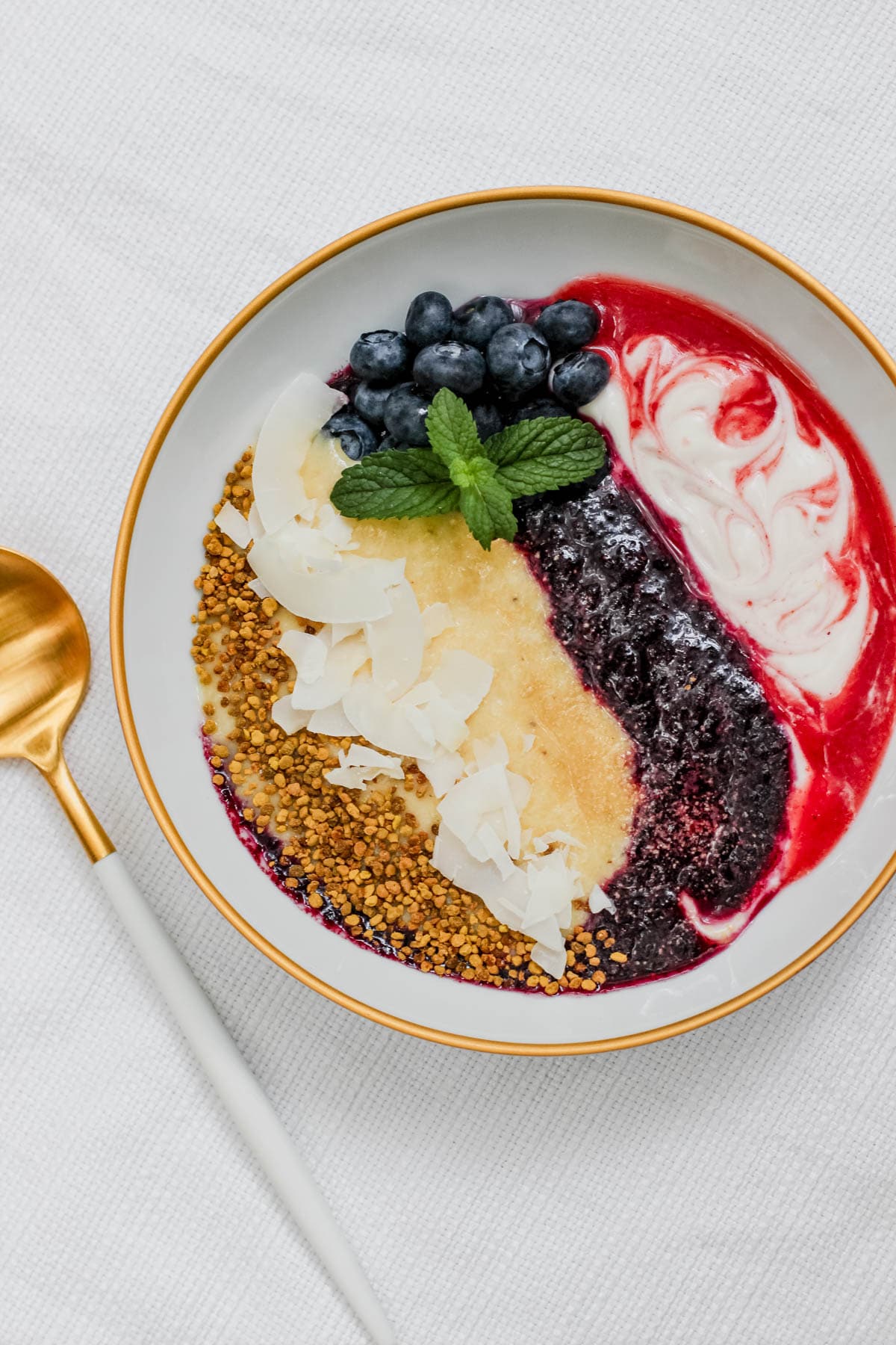 Overhead image of a smoothie yogurt bowl with layered strawberry, blueberry, and pineapple coconut blends, swirled with yogurt and topped with blueberries, bee pollen, coconut, and mint.