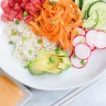 A bright and colorful sushi bowl featuring diced ahi tuna, sliced cucumber, mango, avocado, ribboned carrots, radish, and coconut rice, all drizzled with sriracha aioli.