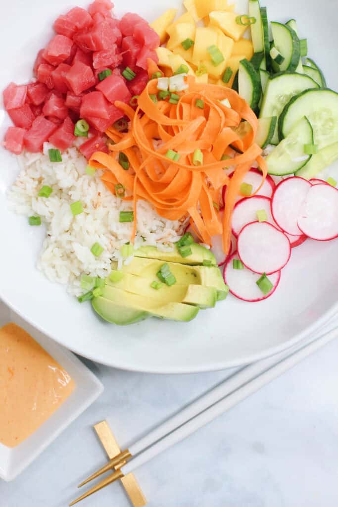 A bright and colorful sushi bowl featuring diced ahi tuna, sliced cucumber, mango, avocado, ribboned carrots, radish, and coconut rice, all drizzled with sriracha aioli.