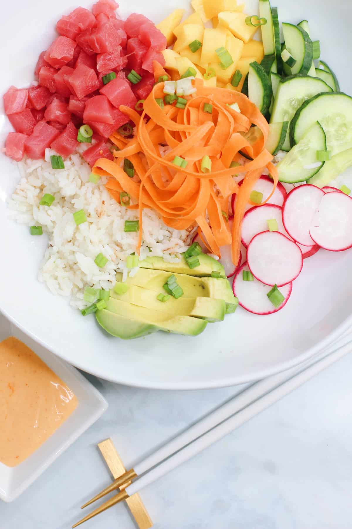 A bright and colorful sushi bowl featuring diced ahi tuna, sliced cucumber, mango, avocado, ribboned carrots, radish, and coconut rice, all drizzled with sriracha aioli.