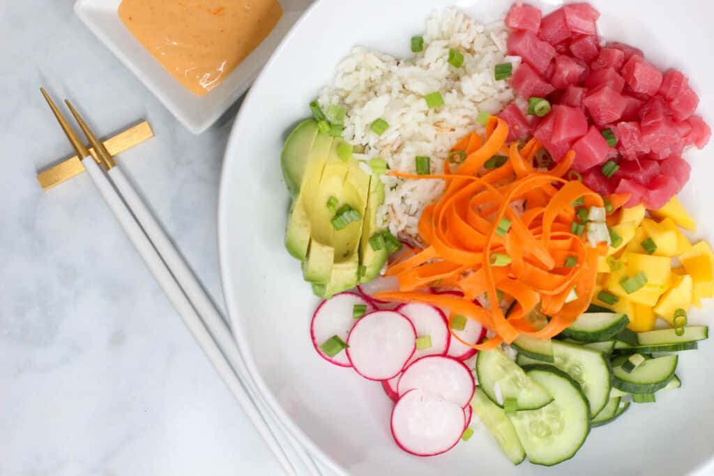 A bright and colorful sushi bowl featuring diced ahi tuna, sliced cucumber, mango, avocado, ribboned carrots, radish, and coconut rice, all drizzled with sriracha aioli.