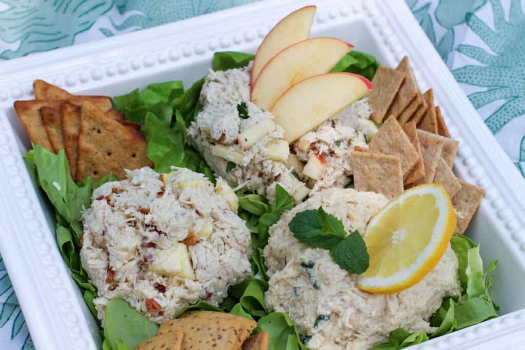 Close up of a chicken salad trio on a white platter, showing creamy texture, all atop a bed of greens served with a variety of crackers.. The platter is displayed on a picnic blanket.