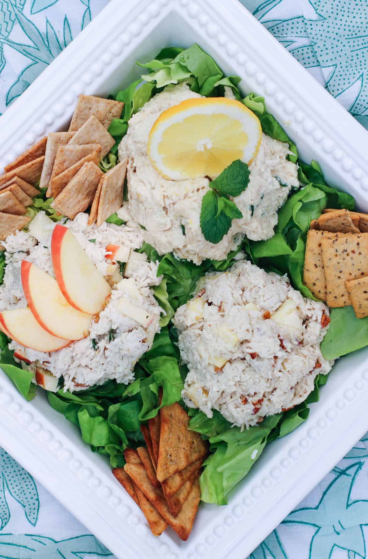 Close up of a chicken salad trio on a white platter, showing creamy texture, all atop a bed of greens served with a variety of crackers.. The platter is displayed on a picnic blanket.