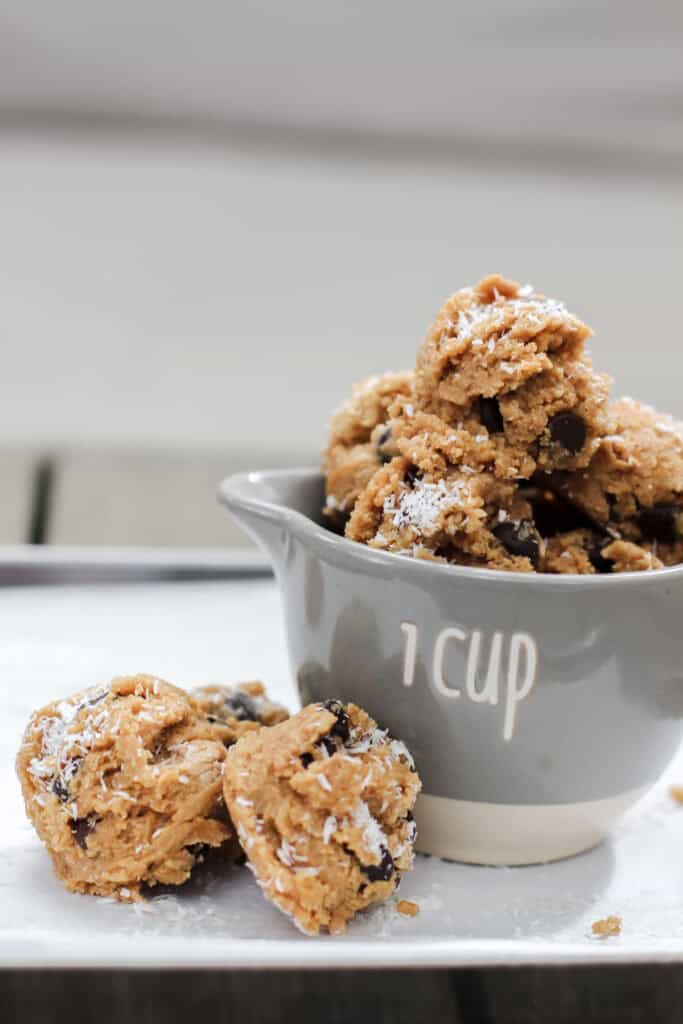 Close-up of coconut chocolate chip protein bites stacked in a 1-cup measuring cup, topped with shredded coconut and chocolate chips, on a white tray.