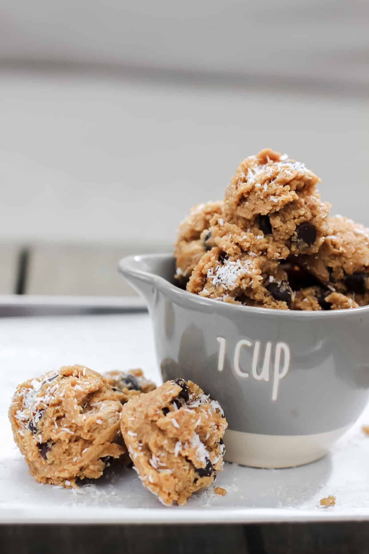 Close-up of coconut chocolate chip protein bites stacked in a 1-cup measuring cup, topped with shredded coconut and chocolate chips, on a white tray.