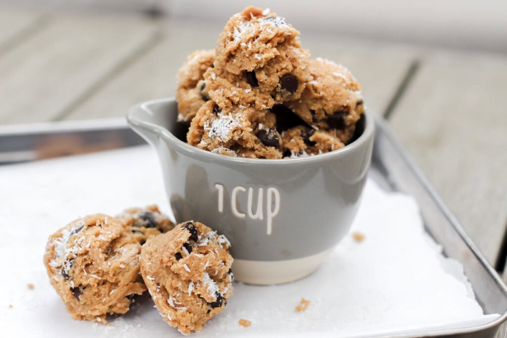 Close-up of coconut chocolate chip protein bites stacked in a 1-cup measuring cup, topped with shredded coconut and chocolate chips, on a white tray.