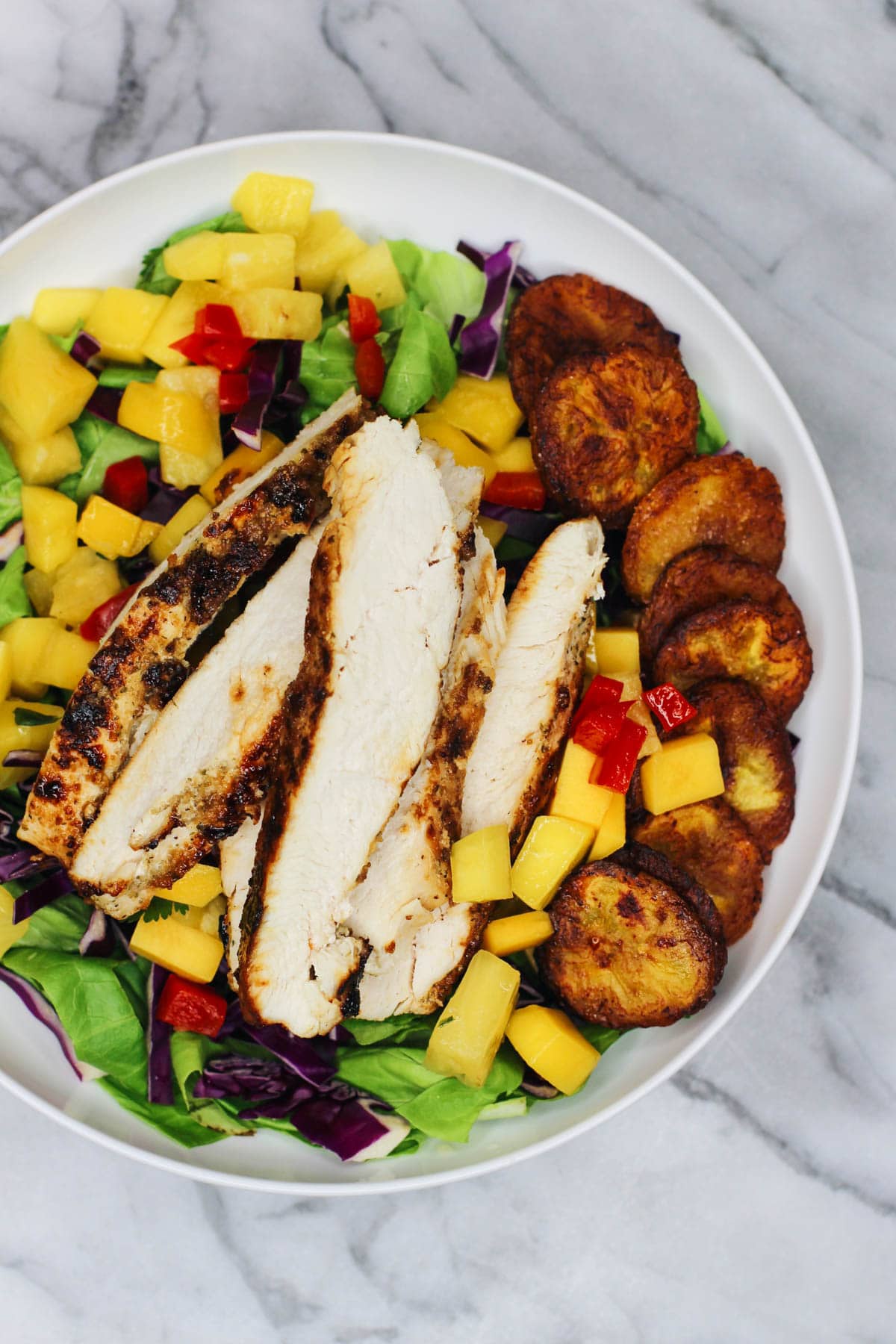 Cuban-inspired salad with grilled chicken, crispy golden plantain “croutons”, and pineapple mango salsa over a bed of lettuce and red cabbage, served in a white bowl on a marble background.
