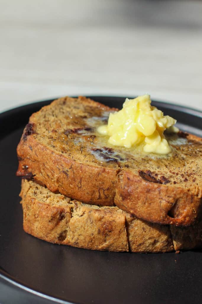 Two thick slices of chocolate chip banana bread topped with melty butter on a matte black plate. The loaf is golden brown, studded with chocolate chips, and perfectly soft and moist.