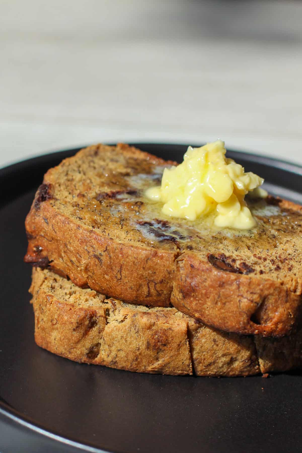 Two thick slices of chocolate chip banana bread topped with melty butter on a matte black plate. The loaf is golden brown, studded with chocolate chips, and perfectly soft and moist.