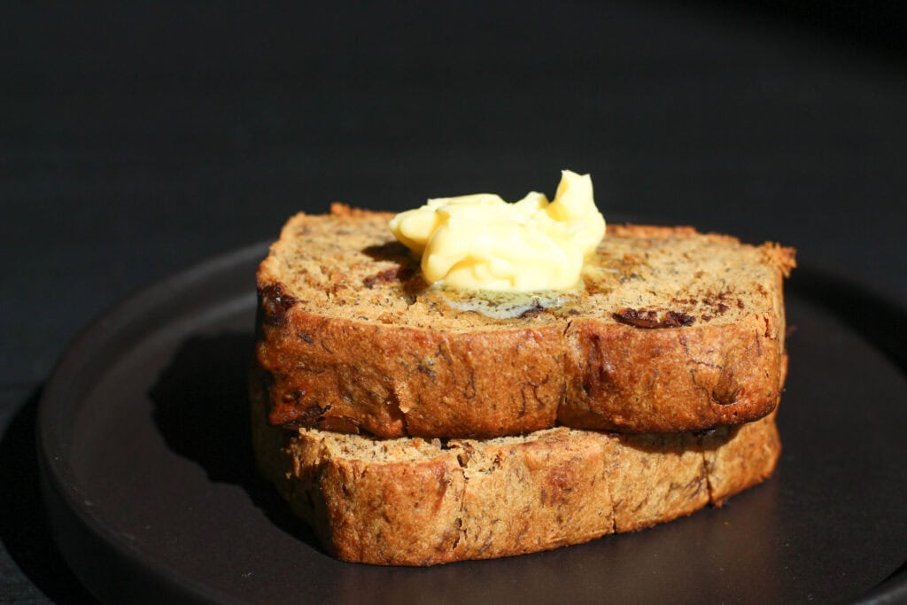 Two thick slices of chocolate chip banana bread topped with melty butter on a matte black plate. The loaf is golden brown, studded with chocolate chips, and perfectly soft and moist.