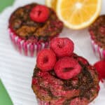 Close-up of three lemon raspberry swirl muffins topped with fresh raspberries, styled on a white plate with lemon slices in the background.