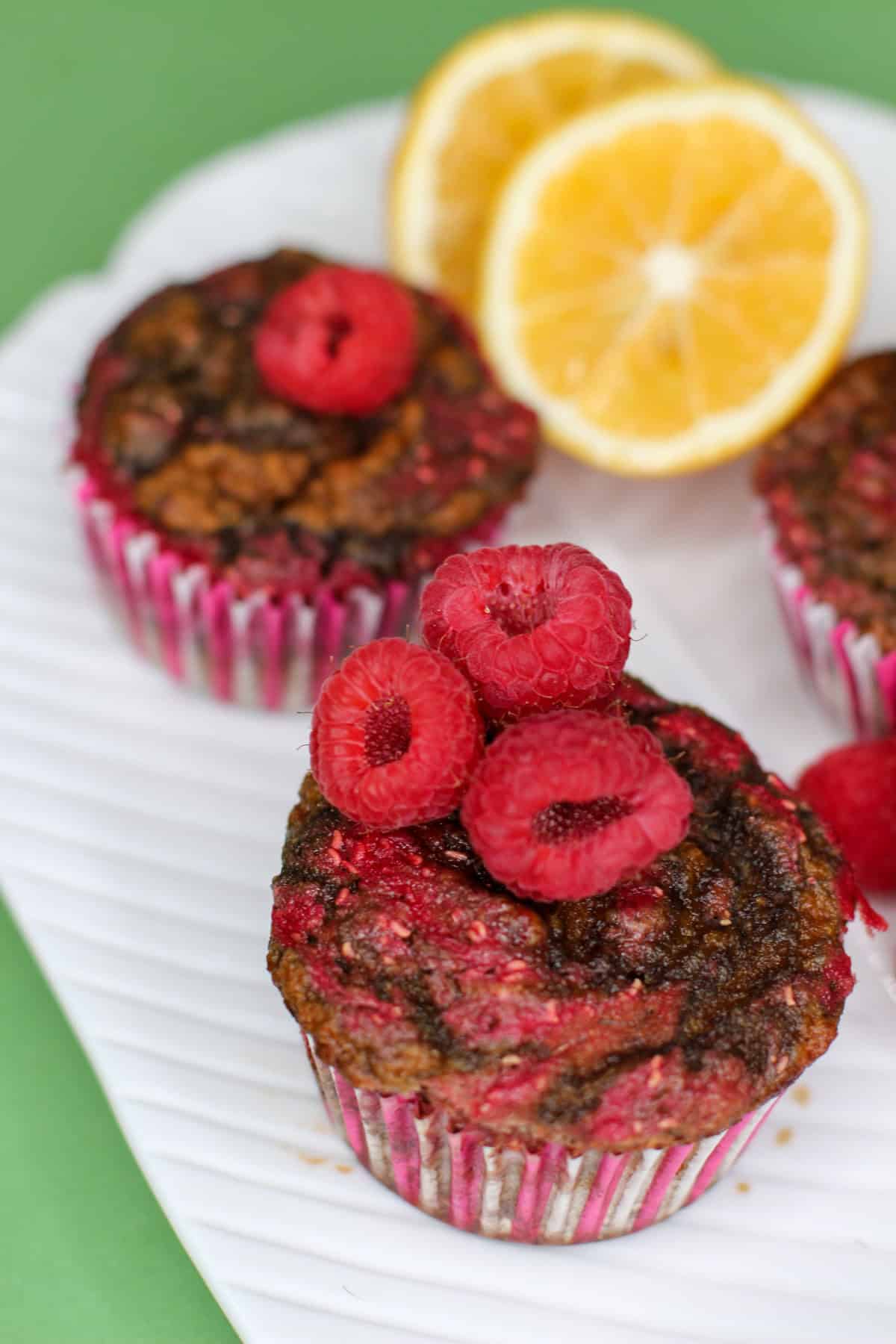 Close-up of three lemon raspberry swirl muffins topped with fresh raspberries, styled on a white plate with lemon slices in the background.