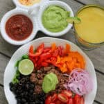 A colorful taco salad bowl with seasoned ground beef, sliced bell peppers, tomatoes, olives, red onion, and a big scoop of avocado dressing. Served alongside a trio of dips and a fresh lime margarita.