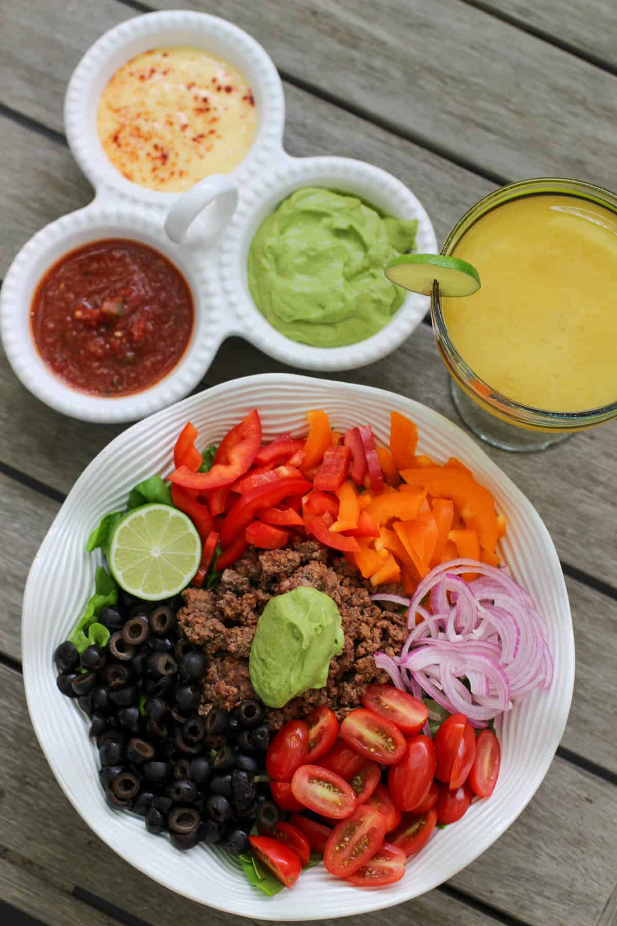A colorful taco salad bowl with seasoned ground beef, sliced bell peppers, tomatoes, olives, red onion, and a big scoop of avocado dressing. Served alongside a trio of dips and a fresh lime margarita.
