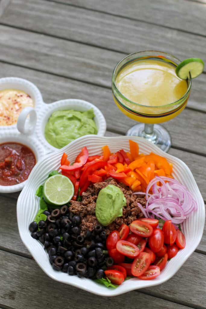 A colorful taco salad bowl with seasoned ground beef, sliced bell peppers, tomatoes, olives, red onion, and a big scoop of avocado dressing. Served alongside a trio of dips and a fresh lime margarita.