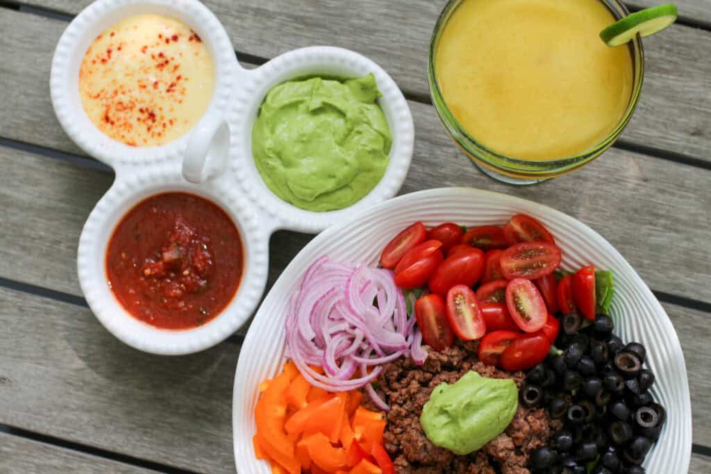 A colorful taco salad bowl with seasoned ground beef, sliced bell peppers, tomatoes, olives, red onion, and a big scoop of avocado dressing. Served alongside a trio of dips and a fresh lime margarita.