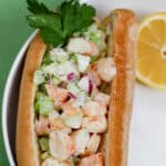 Overhead image of a New England shrimp salad roll served in a gluten-free hot dog bun with chopped celery, onion, and creamy avocado mayo. Garnished with parsley and served with lemon wedge.