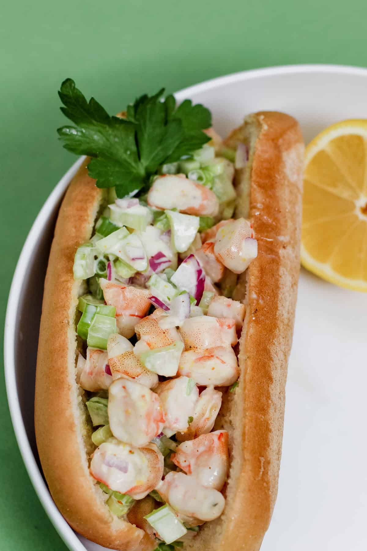 Overhead image of a New England shrimp salad roll served in a gluten-free hot dog bun with chopped celery, onion, and creamy avocado mayo. Garnished with parsley and served with lemon wedge.