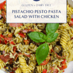 Overhead image of a pesto pasta salad in a white bowl, filled with fusilli pasta, shredded chicken, sliced pepperoni, olives, bell pepper, and topped with fresh basil leaves.