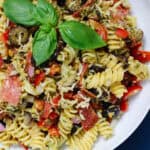 Overhead image of a pesto pasta salad in a white bowl, filled with fusilli pasta, shredded chicken, sliced pepperoni, olives, bell pepper, and topped with fresh basil leaves.