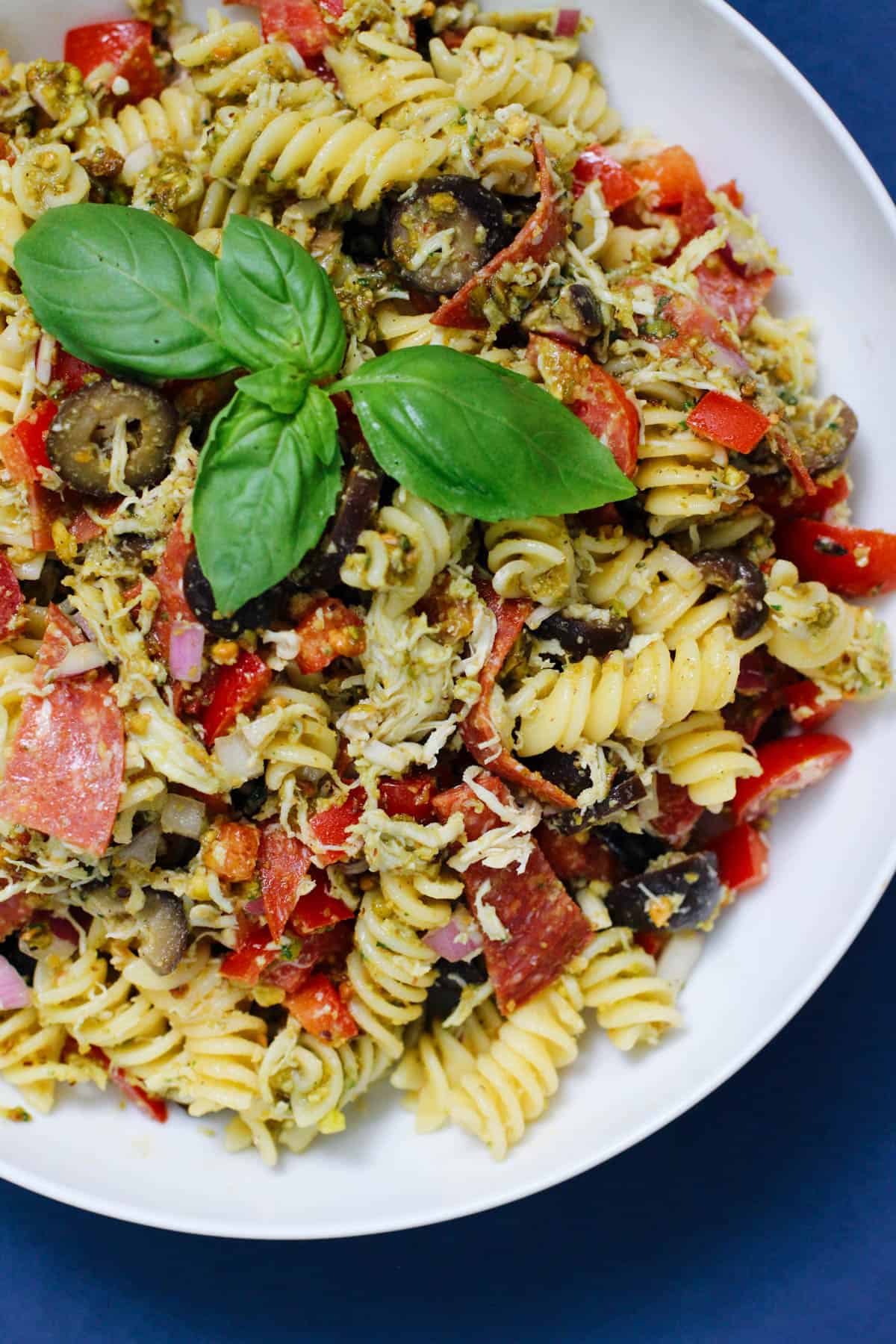 Overhead image of a pesto pasta salad in a white bowl, filled with fusilli pasta, shredded chicken, sliced pepperoni, olives, bell pepper, and topped with fresh basil leaves.