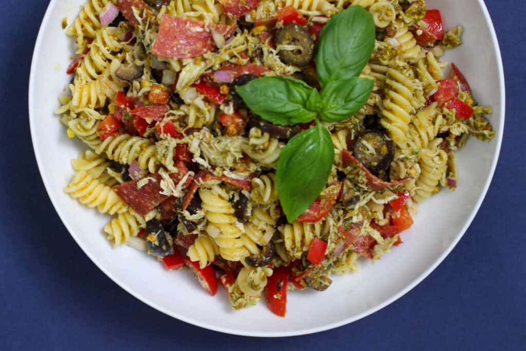 Overhead image of a pesto pasta salad in a white bowl, filled with fusilli pasta, shredded chicken, sliced pepperoni, olives, bell pepper, and topped with fresh basil leaves.