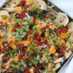 Close-up of baked sheet pan nachos topped with dairy-free queso, sliced black olives, red onion, fresh herbs, and salsa on a silver tray.