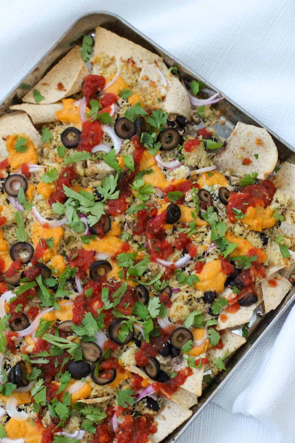 Close-up of baked sheet pan nachos topped with dairy-free queso, sliced black olives, red onion, fresh herbs, and salsa on a silver tray.