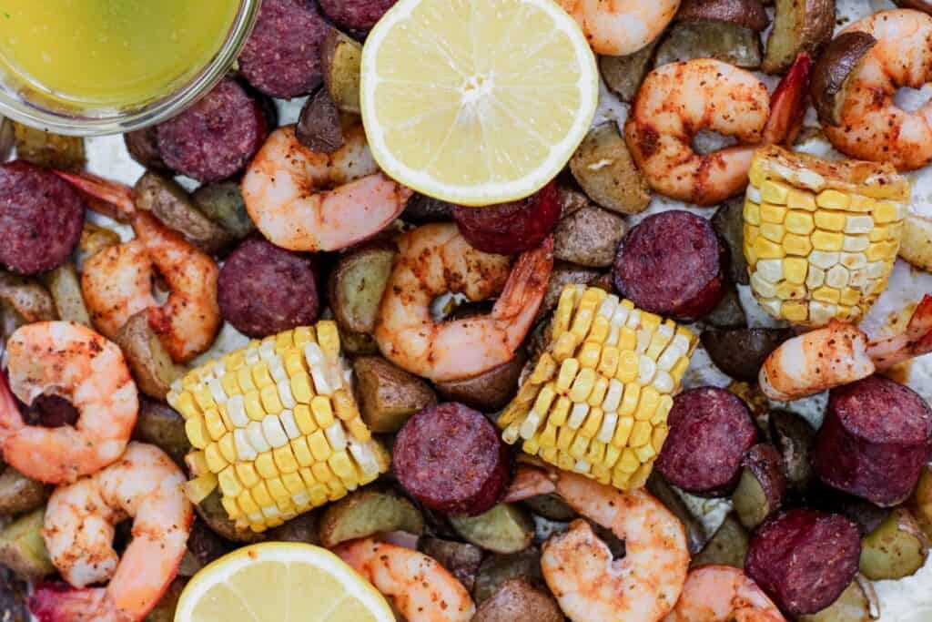 A sheet pan filled with roasted shrimp, sliced andouille sausage, corn on the cob, and baby potatoes. Served with lemon parsley butter and garnished with fresh lemon slices