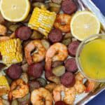 A sheet pan filled with roasted shrimp, sliced andouille sausage, corn on the cob, and baby potatoes. Served with lemon parsley butter and garnished with fresh lemon slices