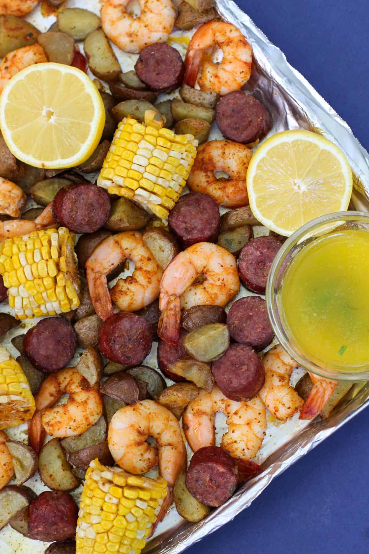 A sheet pan filled with roasted shrimp, sliced andouille sausage, corn on the cob, and baby potatoes. Served with lemon parsley butter and garnished with fresh lemon slices