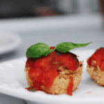 Close-up of a baked Chicken Caprese Muffin Cup topped with warm marinara sauce and a fresh basil leaf, served on a white plate.