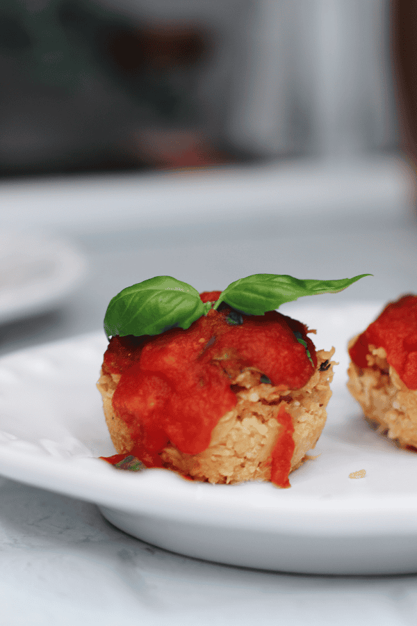 Close-up of a baked Chicken Caprese Muffin Cup topped with warm marinara sauce and a fresh basil leaf, served on a white plate.