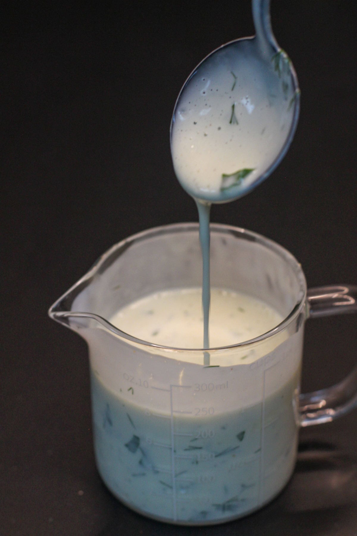 Herbed buttermilk ranch being poured from a spoon into a glass measuring cup.