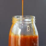 Pouring homemade buffalo sauce with a spoon into a glass measuring cup on a dark countertop.