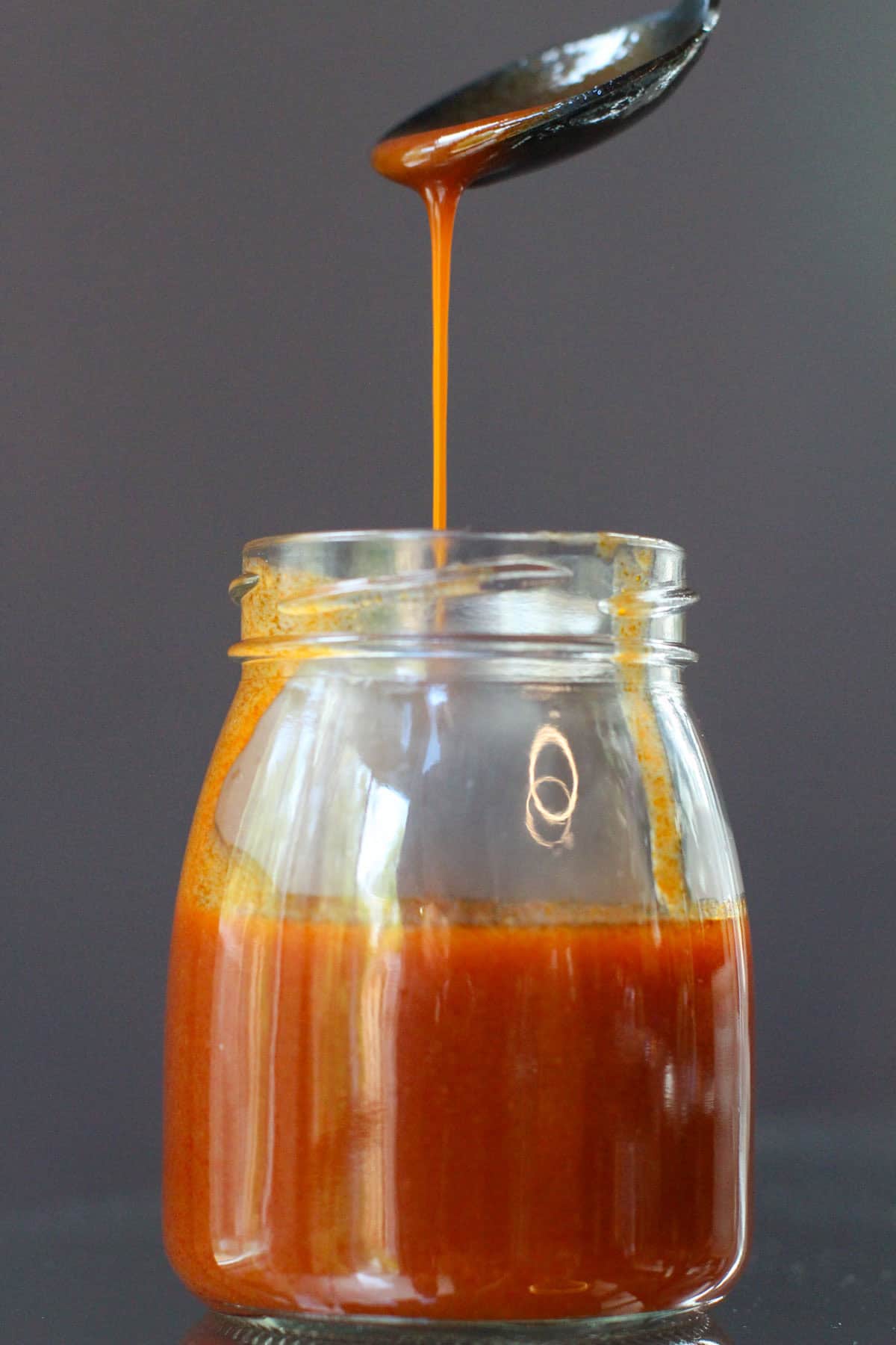 Pouring homemade buffalo sauce with a spoon into a glass measuring cup on a dark countertop.