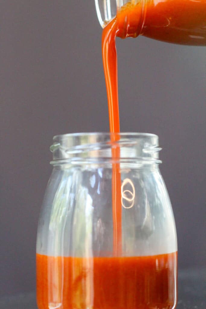 Pouring homemade buffalo sauce with a spoon into a glass measuring cup on a dark countertop.